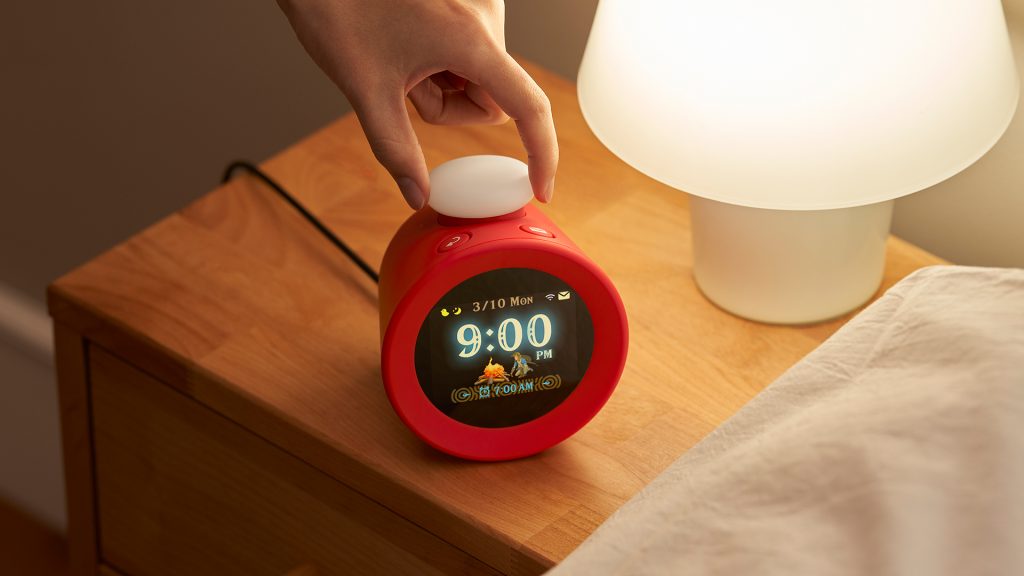 A photograph of Nintendo Sound Clock: Alarmo on a wooden bedside table with a beige/white table lamp.