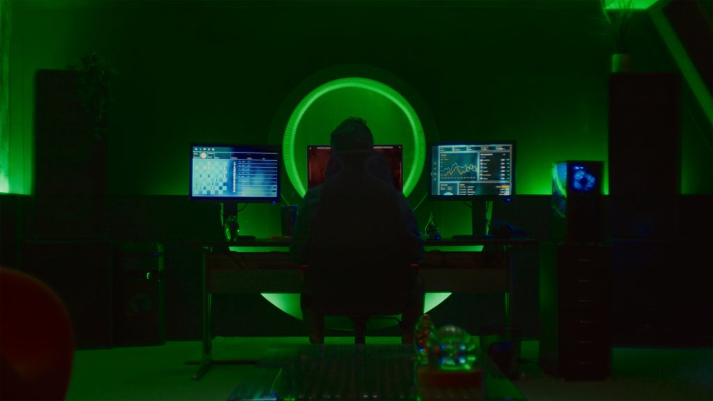 A screenshot from Hello Stranger. A man is sitting in a chair but only his shape can be seen. He is backlit by a large circle of green lights, in front of which are computer monitors.